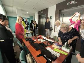 Pokaż zdjęcie: Korytarz, na stolikach leżą sprzęty powiększalniki, lupy, laptop, aparat, telefon, linijka brajlowska. Zebrane osoby oglądają sprzęty.