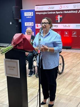 Pokaż zdjęcie: Przy mównicy stoi kobieta w okularach ubrana w niebieską bluzkę. Na mównicy leżą kwiaty.