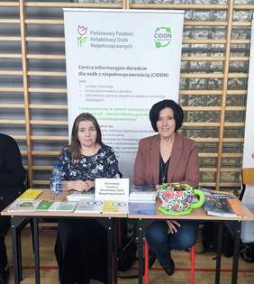 Pokaż zdjęcie: Dwie osoby siedzące za stoiskiem PFRON, przed nimi ułożono broszury, ulotki i materiały informacyjne. W tle widoczny roll‑up z opisem działania Centrów Informacyjno‑Doradczych dla Osób z Niepełnosprawnością.