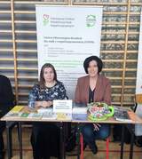 Dwie osoby siedzące za stoiskiem PFRON, przed nimi ułożono broszury, ulotki i materiały informacyjne. W tle widoczny roll‑up z opisem działania Centrów Informacyjno‑Doradczych dla Osób z Niepełnosprawnością.