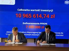 Pokaż zdjęcie: Dwaj mężczyźni podpisują umowę przy stole w sali Pomorskiego Urzędu Wojewódzkiego; w tle ekran z napisem „Całkowita wartość inwestycji 10 965 614,74 zł” oraz „Dofinansowanie PFRON 7 500 000 zł”. Widać logotypy PFRON i urzędu wojewódzkiego.  