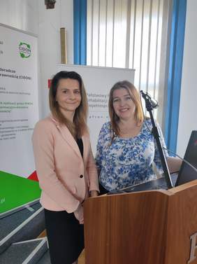 Pokaż zdjęcie: Przy mównicy stoją dwie kobiety skierowane lekko w swoją stronę. Z lewej kobieta w łososiowej marynarce, po prawej kobieta w biało-niebieskiej bluzce. Obydwie się uśmiechają do kamery.