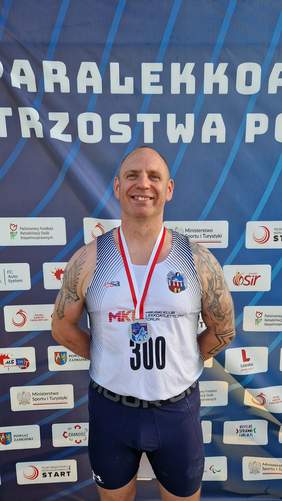 Pokaż zdjęcie: Zawodnik z medalem powieszonym na szyi pozuje do zdjęcia na tle ścianki z logo sponsorów 54. Paralekkoatletycznych Mistrzostw Polski