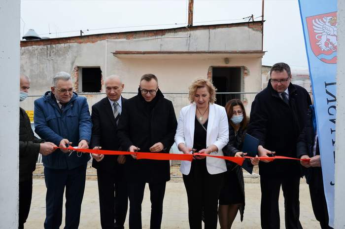 Pokaż zdjęcie: Czterech mężczyzn i dwie kobiety stoją z nożyczkami w ręku przed czerwoną wstęgą, którą będą przecinać. Za nimi stary budynek