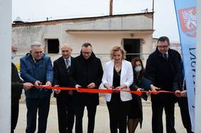 Pokaż zdjęcie: Czterech mężczyzn i dwie kobiety stoją z nożyczkami w ręku przed czerwoną wstęgą, którą będą przecinać. Za nimi stary budynek