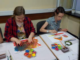 Pokaż zdjęcie: Na zdjęciu dwie młode dziewczyny siedzą przy stole i malują farbami. Uczestniczka warsztatów siedząca po lewej stronie maluje palcami, druga pędzlem. 