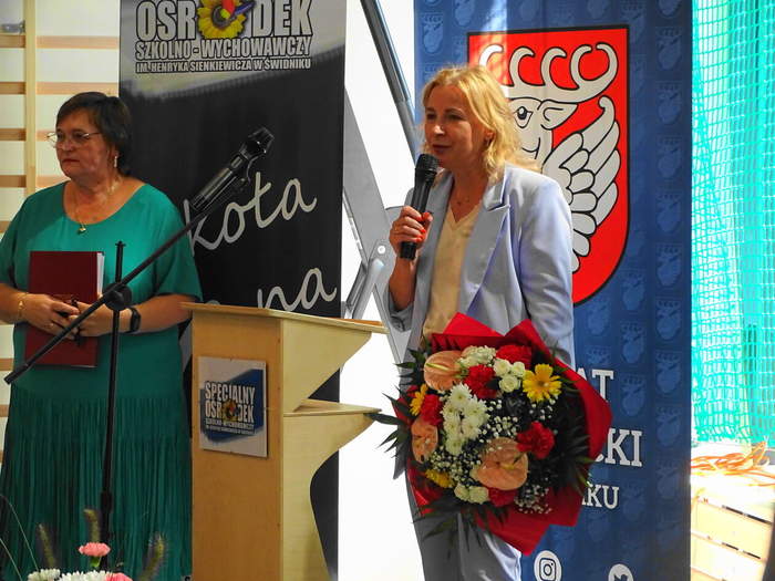 Pokaż zdjęcie: Przy mównicy stoją dwie kobiety. Jedna z nich ubrana w liliowy garnitur trzyma w rękach mikrofon oraz kwiaty.