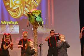 Pokaż zdjęcie: Dzieci w wieku wczesnoszkolnym, ubrane w ciemne koszulki i spodenki w paski, dziewczynki mają warkocze. W trakcie tańca na scenie