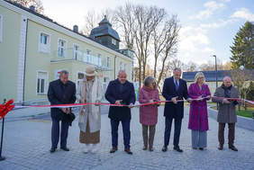 Zdjęcie z ceremonii przecięcia czerwonej wstęgi przed odnowionym, jasnym budynkiem. Siedem osób stoi w rzędzie, każda z nożyczkami, tnąc biało-czerwoną wstęgę. W tle widoczny jest dwupiętrowy budynek w kolorze kremowo-żółtym z balkonem na piętrze i dużym, mansardowym dachem.