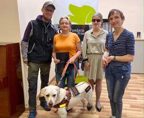 Pokaż zdjęcie: Na tle roll-up'u z napisem "Fundacja Pies Przewodnik" stoi mężczyzna i trzy kobiety. Przed nimi na podłodze stoi pies rasy labrador w kolorze biszkopt.