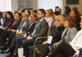 Ujęcie przedstawia pierwszy rząd uczestników konferencji siedzących na szarych krzesłach z drewnianymi podłokietnikami. Wśród nich znajduje się mężczyzna w ciemnoniebieskim garniturze oraz kobieta w białej bluzce z falbaną. Większość osób patrzy w prawą stronę (z perspektywy widza), śledząc prezentację lub wystąpienie. Pod krzesłami widoczne są białe torby konferencyjne z logo „SCWEW”. W tle widać pozostałych uczestników oraz fragment okna, przez które wpada dzienne światło.