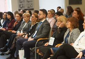 Pokaż zdjęcie: Ujęcie przedstawia pierwszy rząd uczestników konferencji siedzących na szarych krzesłach z drewnianymi podłokietnikami. Wśród nich znajduje się mężczyzna w ciemnoniebieskim garniturze oraz kobieta w białej bluzce z falbaną. Większość osób patrzy w prawą stronę (z perspektywy widza), śledząc prezentację lub wystąpienie. Pod krzesłami widoczne są białe torby konferencyjne z logo „SCWEW”. W tle widać pozostałych uczestników oraz fragment okna, przez które wpada dzienne światło.