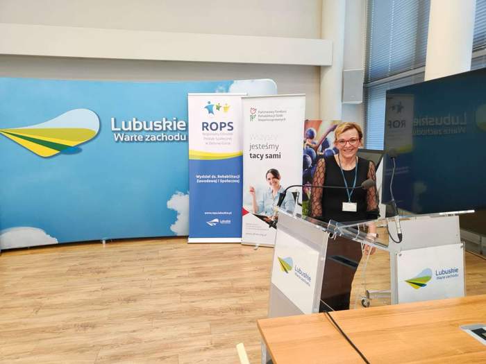 Pokaż zdjęcie: Uśmiechnięta kobieta, która jest ubrana w czarym kolorze, na tle banerów PFRON i Urzędu Marszałkowskiego Wojewózdztwa Lubuskiego