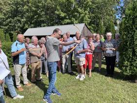 Pokaż zdjęcie: Mężczyzna w koszulce w paski strzela z łuku, otoczony jest przez grupę obserwujących go ludzi. W tle znajdują się drzewa i drewniany budynek.