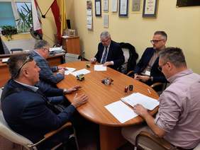 Pokaż zdjęcie: Przy stole siedzi pięciu mężczyzn. Rozmawiają ze sobą. Dwóch panów podpisuje dokumenty. Na stole leżą umowy, pieczątki, telefon, długopisy. 