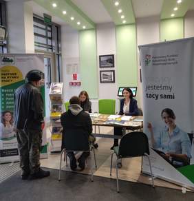 Pokaż zdjęcie: Zdjęcie przedstawia punkt informacyjny, gdzie pracownicy Oddziału Podlaskiego PFRON i Powiatowego Urzędu Pracy w Białymstoku udzielają indywidualnych konsultacji uczestnikom wydarzenia. Na pierwszym planie znajdują się osoby siedzące przy stole z materiałami informacyjnymi, w tle widoczne są banery promujące usługi obu instytucji.