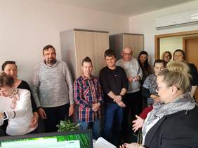 Pokaż zdjęcie: Grupa około jedenastu osób stoi w pomieszczeniu biurowym, ustawiona wzdłuż ściany i szafek. Na pierwszym planie po prawej stronie widać kobietę w okularach, która jest zwrócona w stronę grupy i opowiada o swojej pracy.