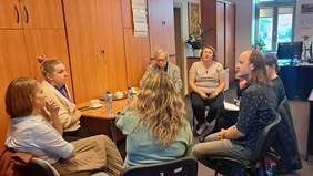 Pokaż zdjęcie: W biurze na krzesłach ustawionych w krąg siedzi kilka osób i rozmawia ze sobą. Po lewej stronie drewniane szafy, w prawym roku okno i biurko na którym stoi komputer.