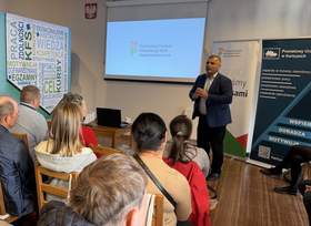 Mężczyzna w garniturze stoi przed grupą osób siedzących na krzesłach. Za nim znajduje się ekran z logo PFRON oraz stoją banery informacyjne. Uczestnicy są zwróceni w stronę prowadzącego.