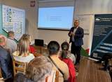 Mężczyzna w garniturze stoi przed grupą osób siedzących na krzesłach. Za nim znajduje się ekran z logo PFRON oraz stoją banery informacyjne. Uczestnicy są zwróceni w stronę prowadzącego.