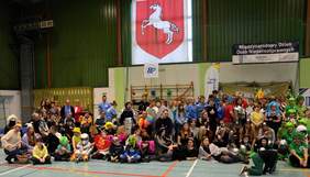 Pokaż zdjęcie: Grupa kilkudziesięciu osób w barwnych strojach pozuje do zdjęcia. Osoby te znajdują się w hali widowiskowo-sportowej. W tle widoczny jest zarówno napis: „Międzynarodowy Dzień Osób Niepełnosprawnych”, jak i logotypy organizatorów wydarzenia.