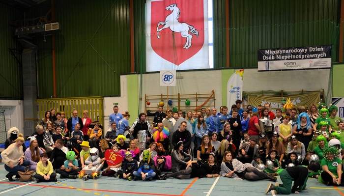 Pokaż zdjęcie: Grupa kilkudziesięciu osób w barwnych strojach pozuje do zdjęcia. Osoby te znajdują się w hali widowiskowo-sportowej. W tle widoczny jest zarówno napis: „Międzynarodowy Dzień Osób Niepełnosprawnych”, jak i logotypy organizatorów wydarzenia.