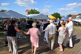 Pokaż zdjęcie: Grupa kilkunastu seniorów stoi w kółku, na drewnianym podeście, trzymając się za ręce. W tle znajdują się ustawione pod namiotami ławki, jak również stoiska poszczególnych wystawców. W oddali znajdują się zabudowania mieszkalne.
