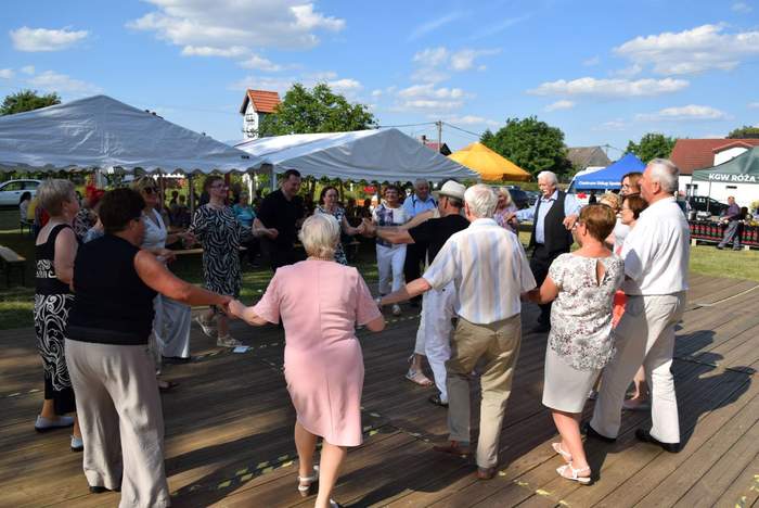 Pokaż zdjęcie: Grupa kilkunastu seniorów stoi w kółku, na drewnianym podeście, trzymając się za ręce. W tle znajdują się ustawione pod namiotami ławki, jak również stoiska poszczególnych wystawców. W oddali znajdują się zabudowania mieszkalne.