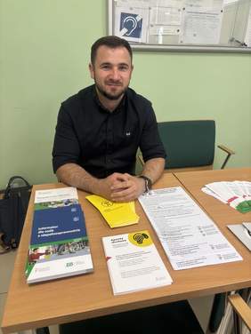 Pokaż zdjęcie: Uśmiechnięty mężczyzna w ciemnej koszuli siedzi przy stole, na którym wyłożone są liczne ulotki oraz informatory.