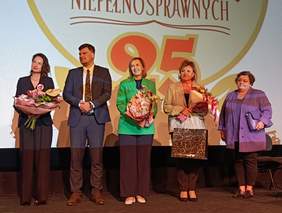 Pokaż zdjęcie: Cztery Kobiety i jeden mężczyzna stoją na scenie, za nimi na ekranie logo Uniwersytetu Trzeciego Wieku i Osób Niepełnosprawnych