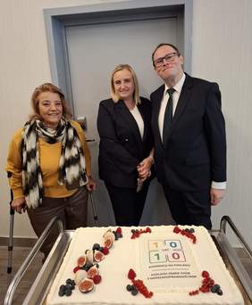 Pokaż zdjęcie: Tort z okazji 10 jubileuszowy Podlaski Konwent Osób z Niepełnosprawnościami. Z tyłu stoją dwie kobiety i mężczyzna. Pierwsza z kobiet opiera się o kule, ubrana w sweter o musztardowym kolorze oraz dopasowane ciemno brązowe, skórzane spodnie z cienkim szalem na szyi. Druga z kobiet blądynka ubrana w czarny garnitur i biała koszulowa bluzkę, trzyma w ręku telefon komórkowy. Mężczyzna w okularach ubrany w czarny garnitur, białą koszulę i czarny krawar.