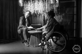 Pokaż zdjęcie: Czarno-białe zdjęcie przedstawiające scenę z Dyrektorem Oddziału i aktorem, z tego samego przedstawienia. Mężczyzna w garniturze i krawacie siedzi po lewej stronie. Po prawej, na wózku inwalidzkim, siedzi kobieta w kimonie. Oboje nachylają się do siebie, a ich dłonie dotykają się na okrągłym, ażurowym stoliku stojącym między nimi. W tle, pod białymi lampionami, wiszą białe girlandy kwiatów.