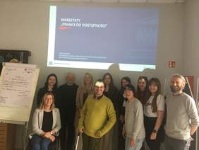 Pokaż zdjęcie: Grupa uśmiechniętych osób pozuje do zdjęcia w sali z projektorem wyświetlającym napis "WARSZTATY 'PRAWO DO DOSTĘPNOŚCI'" i flipchartem z notatkami.