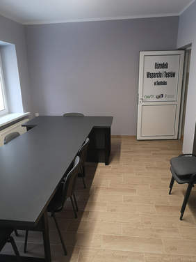 Pokaż zdjęcie: Duży jasny szary pokój, na środku stoją dwa czarne stoły połączone w kształt litery T, przy stole stoją krzesła, podłoga wyłożona brązowymi drewnopodobnymi płytkami gresowymi