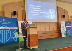 Pokaż zdjęcie: Mężczyzna w garniturze przemawia przy mównicy podczas konferencji w sali wykładowej. Na ekranie za nim widoczny jest slajd z tytułem prezentacji: 'Dyskryminacja pracowników ze względu na niepełnosprawność'. W tle stoją roll-upy z logo Państwowej Inspekcji Pracy oraz Funduszu Rehabilitacji Osób Niepełnosprawnych.