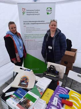 Pokaż zdjęcie: Po dwóch stronach baneru stoją dwie panie. Na banerze zamieszczone są informacje dotyczące PFRON. Przed paniami leżą na stoliku materiały reklamowe i promocyjne. 