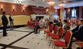 Pokaż zdjęcie: Na zdjęciu uczestnicy spotkania w eleganckiej sali konferencyjnej z czerwonymi krzesłami i dekoracyjnymi ścianami. Kilkanaście osób siedzi, słuchając wystąpienia dwóch prowadzących stojących przed nimi. Trzecia z osób, kobieta trzyma mikrofon, prowadzi dyskusję. Uczestnicy są skoncentrowani na prelegentach.