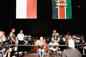 Pokaż zdjęcie: Ring bokserski, na nim dwaj zawodnicy na wózkach inwalidzkich. Jeden z nich trzyma pas mistrzowski. W tle wiszą flagi Polski i Kenii, a wokół ringu znajdują się sędziowie oraz inni uczestnicy Gali.