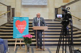 Pokaż zdjęcie: Mężczyzna w garniturze przy mównicy. Przemawia do zgromadzonych. Po jego prawej stronie sztaluga z plakatem. Widnieje na nim czerwone serce na niebieskim tle i tekst informujący o konkursie