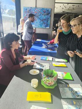 Pokaż zdjęcie: Przy stoliku na którym leżą ulotki siedzi kobieta, przy stoliku stoją osoby zainteresowane ulotkami.