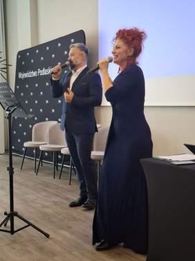 Pokaż zdjęcie: Kobieta i mężczyzna z mikrofonami w ręku otwierają Podlaski Konwent Osób z Niepełnosprawnościami – „Konwencja jest Kobietą”. Mężczyzna z krótką brodą ubrany w granatowy garnitur, białą koszulę oraz czarne pantofle. Kobieta rude włosy spięte w luźny kok, ubrana w długą, lekko dopasowaną suknię dzianinową rozszerzającą się ku dołowi z rękawem trzy czwarte.