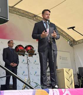 Pokaż zdjęcie: Zbliżenie na scenę podczas oficjalnego wystąpienia. Czterech przedstawicieli instytucji (trzech mężczyzn i jedna kobieta) stoi na tle banerów różnych organizacji, w tym ZUS, Pomorze Zachodnie i Floating Garden Szczecin. Mężczyzna przy mikrofonie przemawia, w tle widać instrumenty muzyczne i artystyczną grafikę.