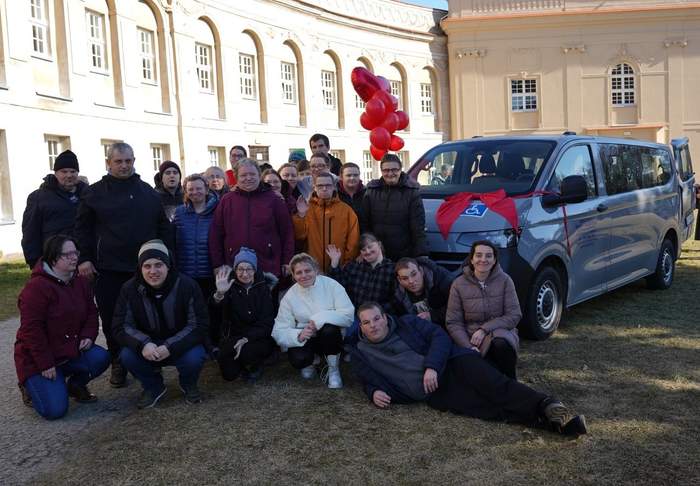 Pokaż zdjęcie: Fotografia przedstawia kilkanaście osób z Warsztatu Terapii Zajęciowej prowadzonego przez Stowarzyszenie PRO-AKTIV, którzy stoją i kucających na trawie przed jasnym, dużym budynkiem z wieloma oknami. Po prawej stronie zdjęcia stoi srebrny bus, ozdobiony dużą czerwoną kokardą na masce. Nad grupą, mniej więcej na środku, widać czerwone balony. Większość osób ma zimowe kurtki i czapki; kilka osób unosi ręce w geście machania. 