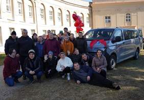Pokaż zdjęcie: Fotografia przedstawia kilkanaście osób z Warsztatu Terapii Zajęciowej prowadzonego przez Stowarzyszenie PRO-AKTIV, którzy stoją i kucających na trawie przed jasnym, dużym budynkiem z wieloma oknami. Po prawej stronie zdjęcia stoi srebrny bus, ozdobiony dużą czerwoną kokardą na masce. Nad grupą, mniej więcej na środku, widać czerwone balony. Większość osób ma zimowe kurtki i czapki; kilka osób unosi ręce w geście machania. 