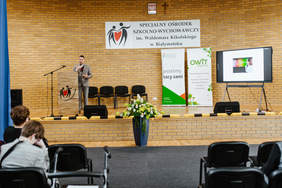 Pokaż zdjęcie: Po prawej mężczyzna w garniturze w dłoni trzyma mirkofon. Za nim wisi baner z napisem Specjalny Ośrodek Szklono-Wychowawczy im. Waldemara Kikolskiego w Białymstoku oraz stoją dwa banery z logo PFRON i OWiT. 