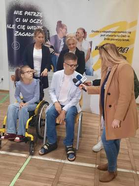 Pokaż zdjęcie: Dwie dziennikarki rozmawiają z chłopcem, który siedzi na krześle. Obok niego jest dziewczynka na wózku inwalidzkim, która uśmiecha się szeroko. 