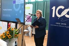 Pokaż zdjęcie: Przy mównicy stoi kobieta w średnim wieku w okularach. W tle widać duży monitor.