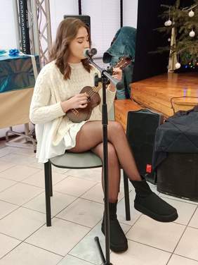Pokaż zdjęcie: Na krzesełku siedzi młoda dziewczyna, która gra na ukulele i śpiewa do mikrofonu. 