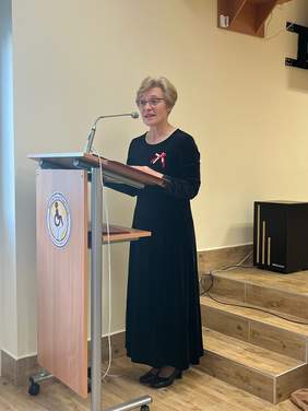 Pokaż zdjęcie: Przy mównicy stoi kobieta ubrana w czarna sukienkę i przemawia do zgromadzonych gości