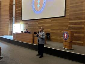 Pokaż zdjęcie: Mężczyzna w jasnoszarej marynarce koszuli i ciemnych spodniach stoi i trzyma w ręku mikrofon. Za nim podwyższenie na którym po prawej stoi mównica po lewej duże biurko za którym siedzi kobieta. Wszystko na tle drewniane ściany na której wisi du